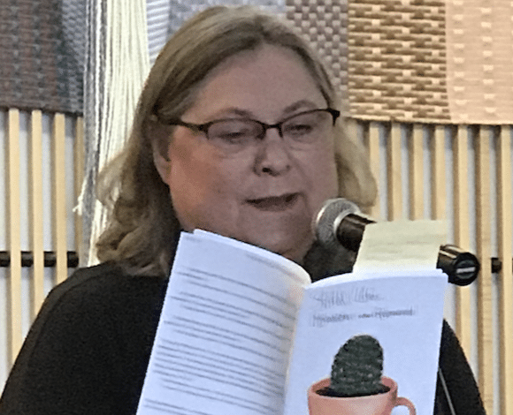 Woman in glasses holds a white book and speaks into a microphone.